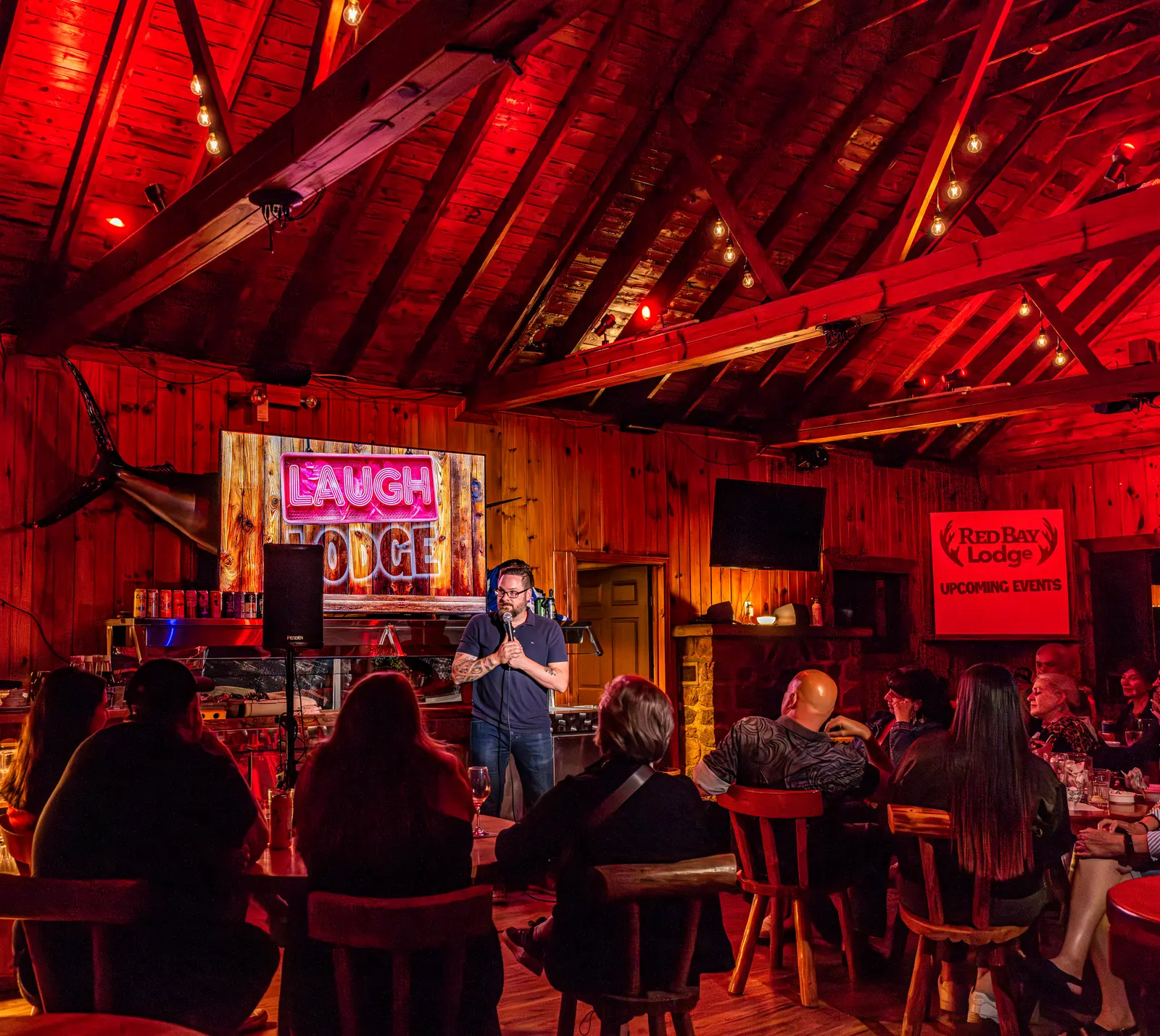A comedian on stage at a Laugh Lodge night in the red-lit lodge, the audience in the foreground