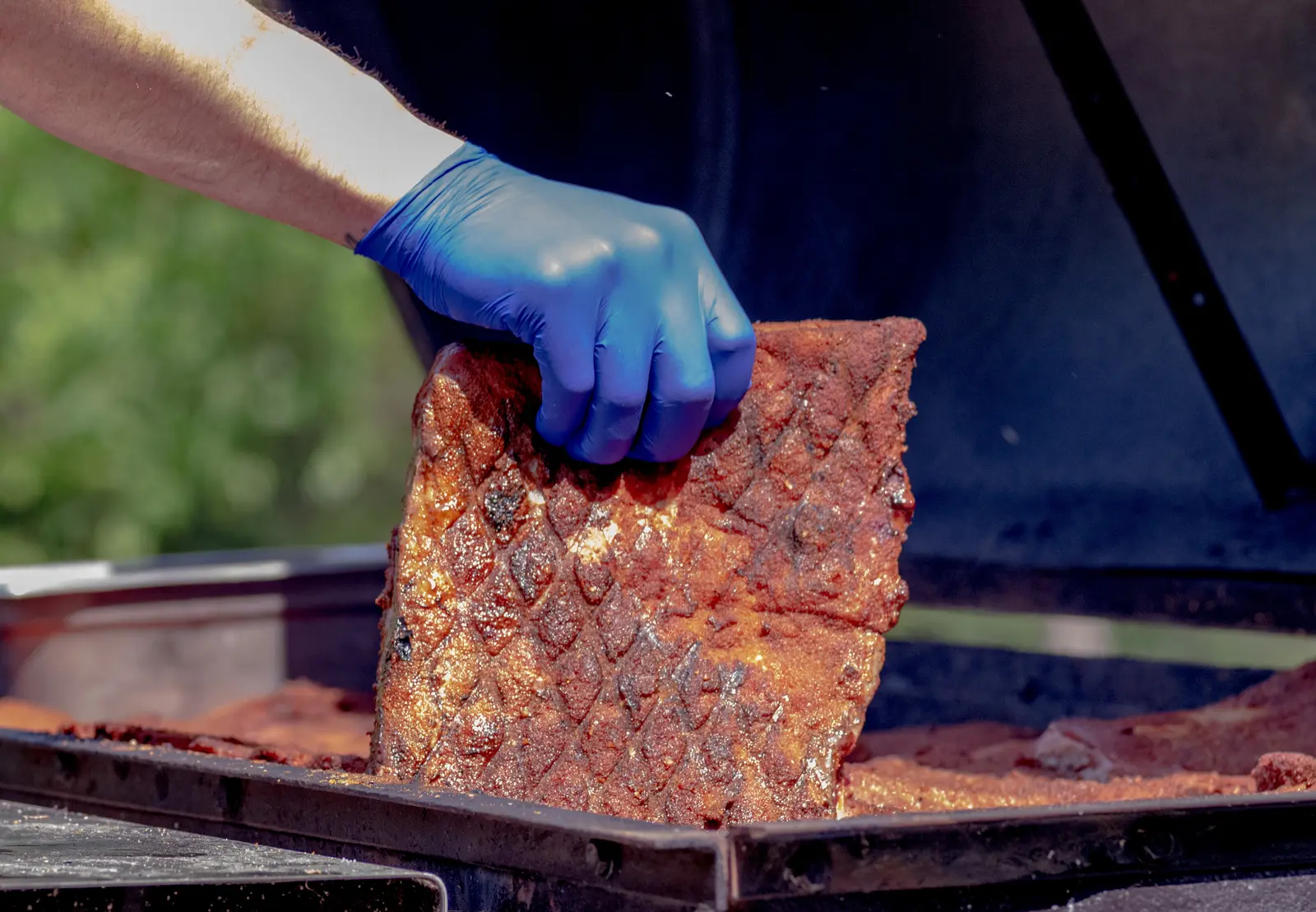 A cook in a blue glove lifting a smoked rack of meat off the tray