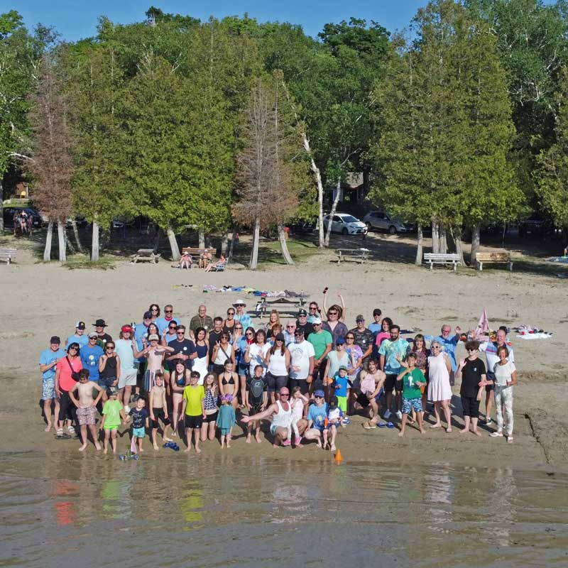 A large group of people gathered on the sand at Red Bay Beach, with cedars behind them