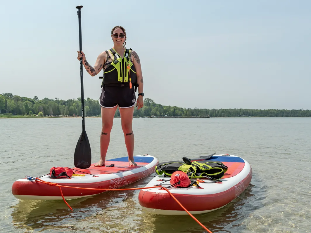 A smiling paddleboarder in a life jacket standing on a board in shallow water, with a second board beside her