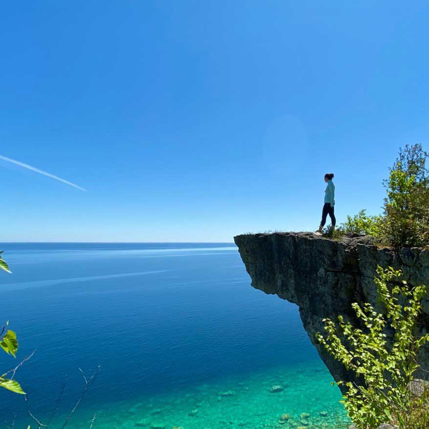 A hiker standing on a limestone cliff edge above clear blue water on the Bruce Peninsula