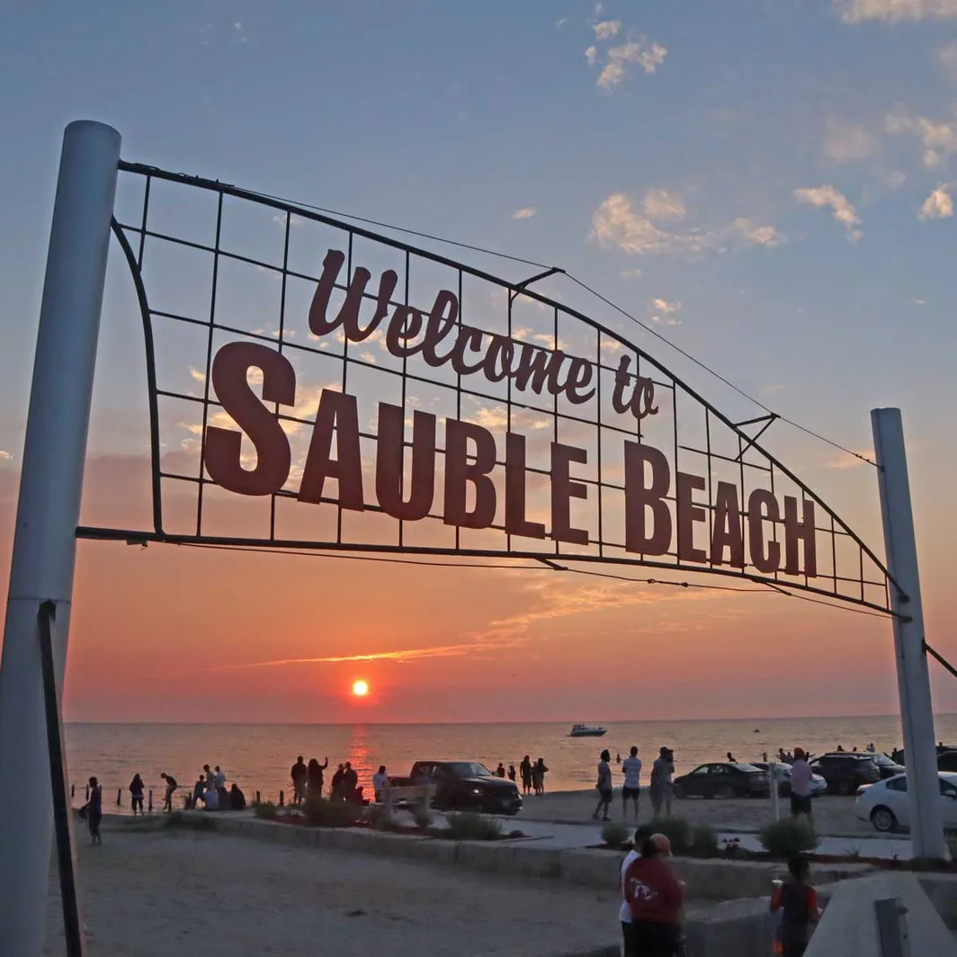 People watching the sun set over Lake Huron beneath the Welcome to Sauble Beach sign
