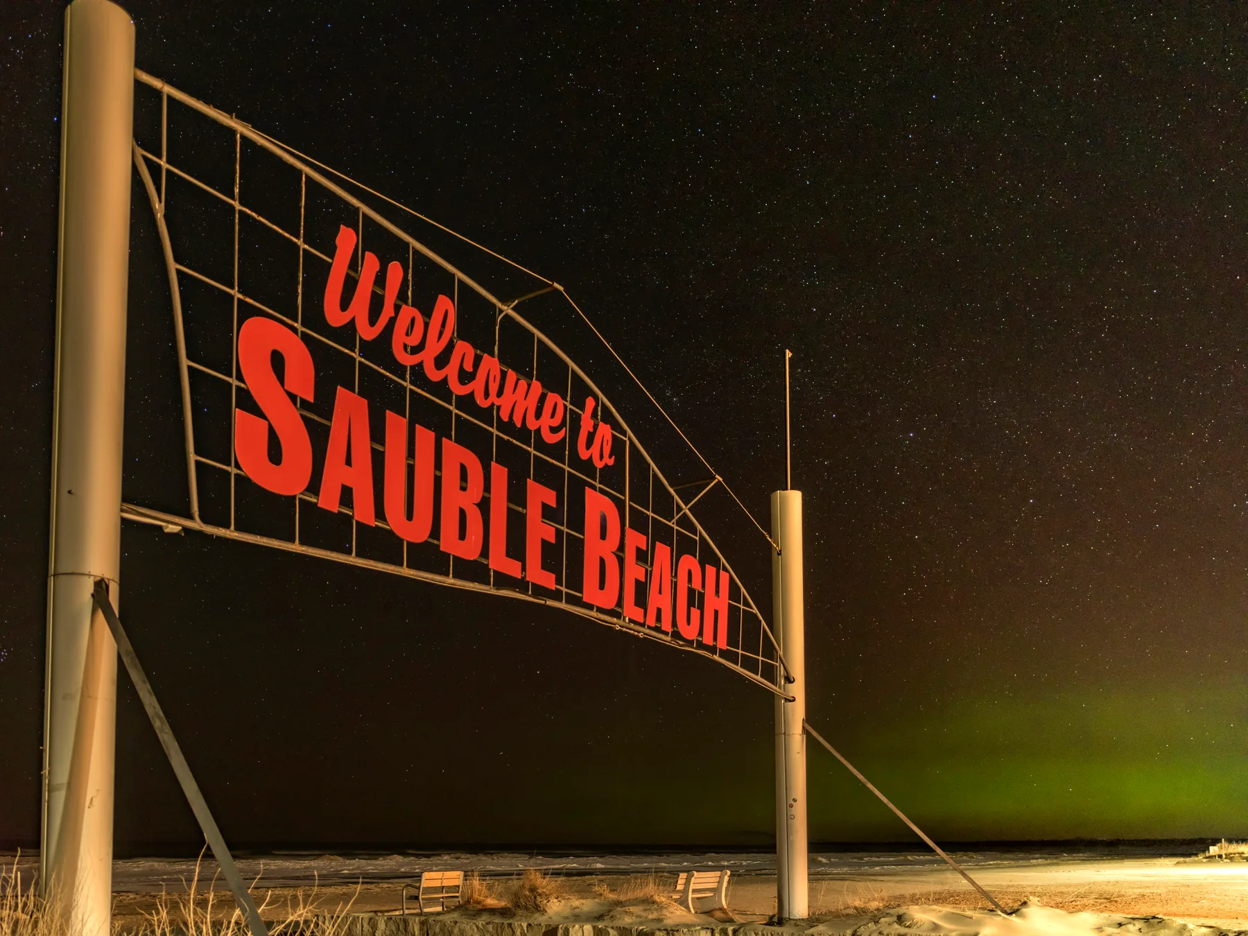 The Welcome to Sauble Beach sign at night under a starry sky with a faint green aurora on the horizon